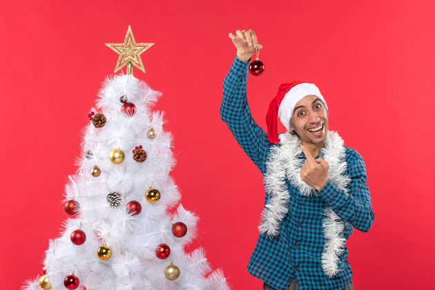 站着一个滑稽的年轻人 戴着圣诞老人帽 穿着一件蓝色的衬衫 站在红色的圣诞树旁 举着一副装饰饰品庆祝帽子年轻人