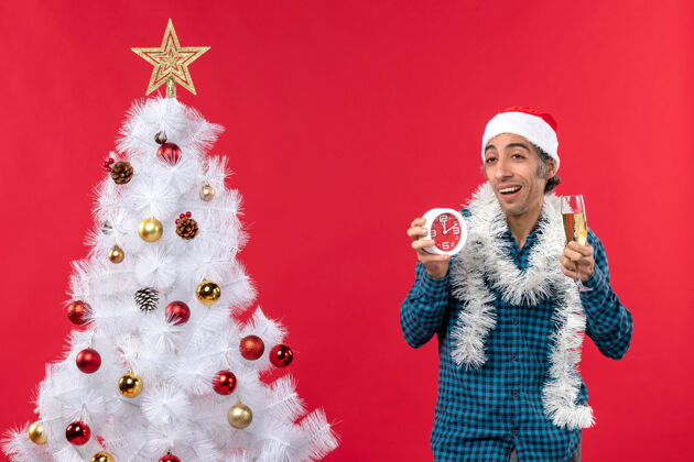 圣诞树不自信的年轻人戴着圣诞老人的帽子 手里拿着一杯葡萄酒 站在红地毯上的圣诞树旁的钟圣诞小伙子小伙子