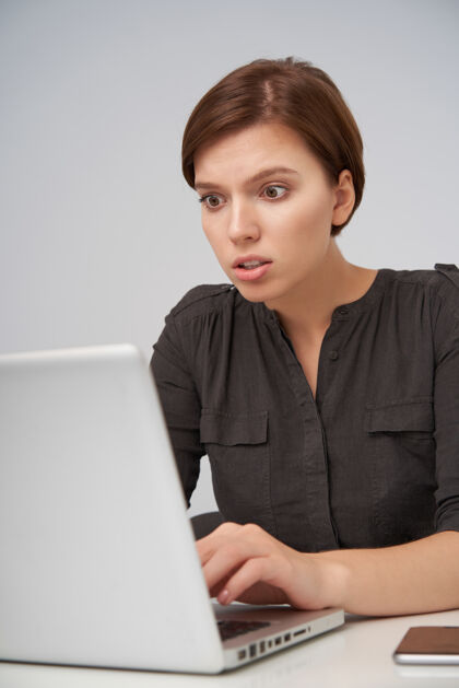黑色困惑的棕色头发的年轻女人 随意的发型 手放在笔记本电脑的键盘上 看着屏幕 困惑的脸 孤立的白色办公室年轻服装