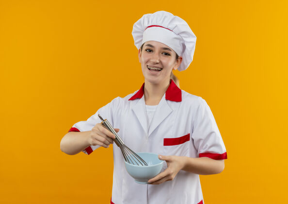 年轻快乐的年轻漂亮厨师 身着厨师制服 戴着牙套 拿着搅拌器和碗 隔离在橙色的空间里橙色制服厨师
