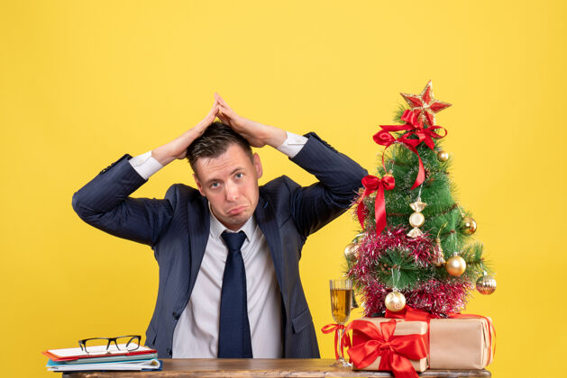 树不快乐的男人 他的手坐在圣诞树附近的桌子和黄色的礼物屋顶房子的正面视图坐着制造快乐