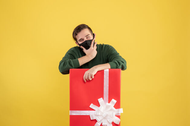 面具正面图：戴着黑色面具的体贴的年轻人站在黄色的大礼盒后面男人礼品盒黑色