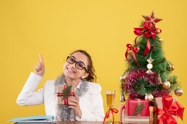 树前视图：女医生拿着圣诞礼物和黄色背景上的圣诞树坐着礼物插花健康