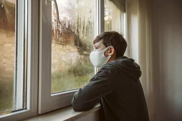 冠状病毒戴着口罩的男孩斜着身子看着窗外预防呆在家里疾病