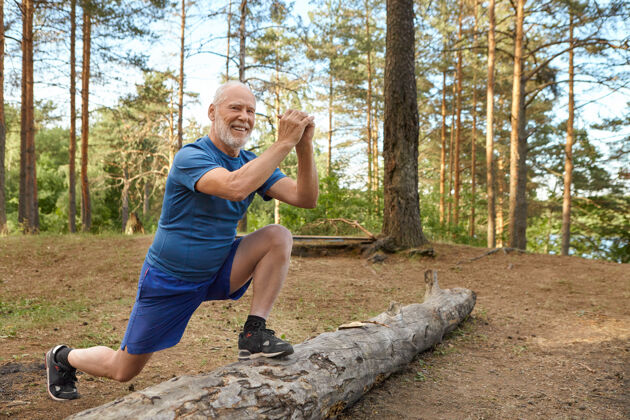 苔藓积极开朗的欧洲男性退休者 穿着t恤 短裤和运动鞋 在户外进行热身活动 一只脚站在原木上 双手放在他面前 做弓箭步 微笑着跑步者脚人