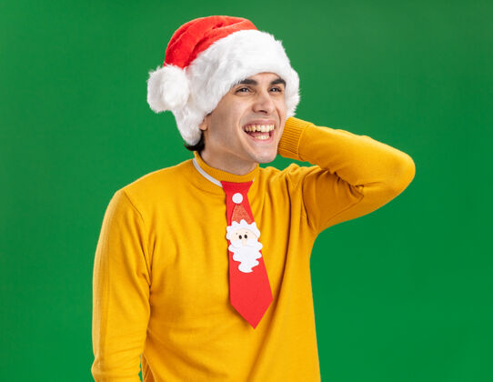 年轻一个穿着黄色高领毛衣 戴着圣诞帽 打着滑稽领带的年轻人站在绿色的背景上 面带幸福的笑容 站在一旁看着圣诞老人帽子微笑