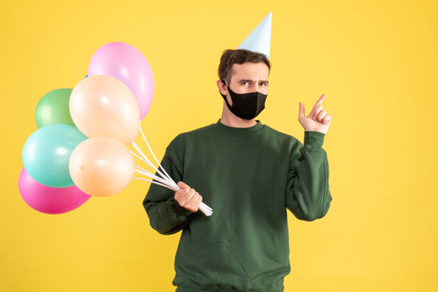 男人正面图：戴着派对帽的年轻人 彩色气球指向后面站在黄色的舞台上气球年轻人聚会