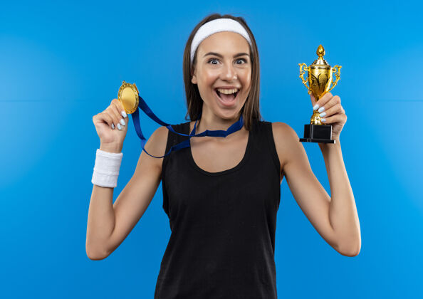 周围快乐的年轻漂亮的运动女孩戴着头带和腕带 脖子上戴着奖牌 拿着冠军杯和奖牌孤立在蓝色的空间里运动蓝色奖牌