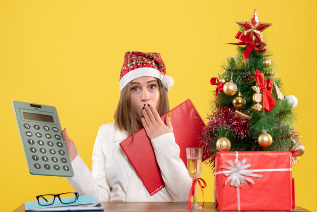 人正面图：女医生拿着圣诞礼物坐在黄色桌子上拿着计算器桌子圣诞节微笑