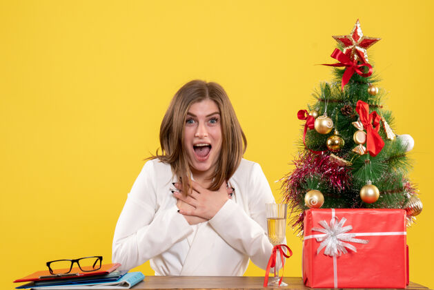 背景正面图：女医生坐在黄色背景的桌子前 放着圣诞树和礼品盒快乐坐着女性