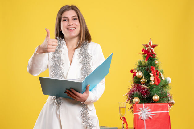 快乐正面图：女医生站着拿着文件放在黄色的桌子上 桌子上放着圣诞树和礼品盒成人女医生前面