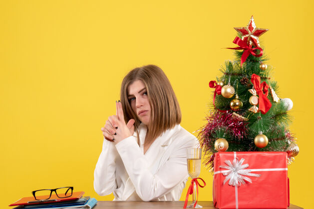 快乐正面图：女医生坐在黄色背景的桌子前 放着圣诞树和礼品盒人办公室圣诞节