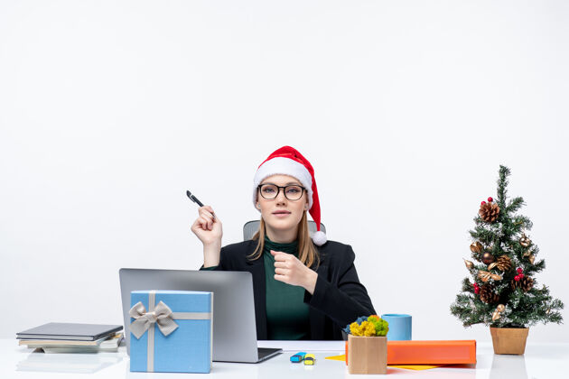 公司戴着圣诞老人帽的金发女人坐在桌子旁 手里拿着圣诞树和礼物圣诞节圣诞老人圣诞树