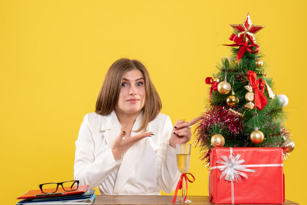 新的正面图：女医生坐在黄色背景的桌子前 放着圣诞树和礼品盒微笑医生主任