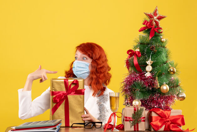 病毒前视图女医生戴着面具坐着 圣诞礼物和黄色背景上的树礼物医生新