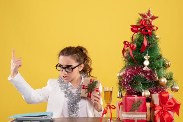 女医生前视图：女医生拿着圣诞礼物和黄色背景上的圣诞树坐着情感成人礼物