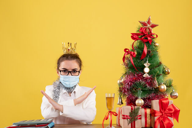 成人正面图女医生戴着黄色背景的无菌口罩坐在圣诞树和礼盒里健康人医生