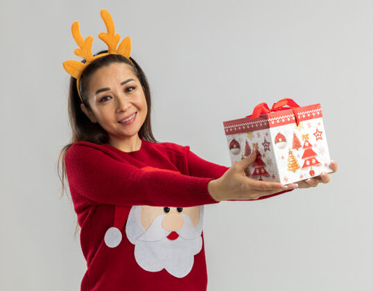 毛衣穿着红色圣诞毛衣的年轻女子 戴着滑稽的鹿角边 手里拿着圣诞礼物 脸上洋溢着快乐和积极的笑容站着拿着鹿