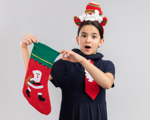 边小女孩穿着针织连衣裙 头上系着一条红色领带 头上戴着一个有趣的圣诞边 手里拿着圣诞长袜 看起来很惊讶长袜站着新
