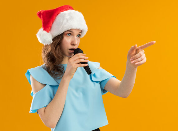边穿着蓝色上衣 戴着圣诞帽的严肃的年轻女子对着麦克风说话 食指指向一边新年轻严肃