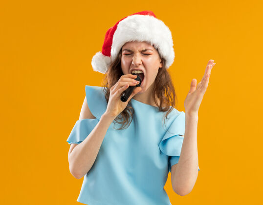 橙色身穿蓝色上衣 头戴圣诞帽的年轻女子举起手臂对着麦克风大喊大叫 激动得发狂年麦克风顶部