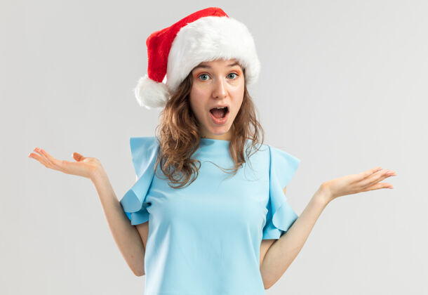 新的戴着蓝色上衣和圣诞帽的年轻女子举起双臂 看上去既困惑又惊讶帽子站着困惑