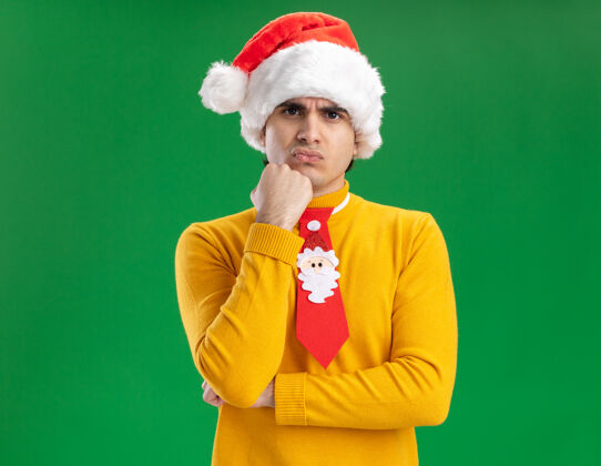 领带一个穿着黄色高领毛衣 戴着圣诞帽 打着滑稽领带的年轻人看着摄像机 不高兴地用手托着下巴站在绿色背景上年轻站着手