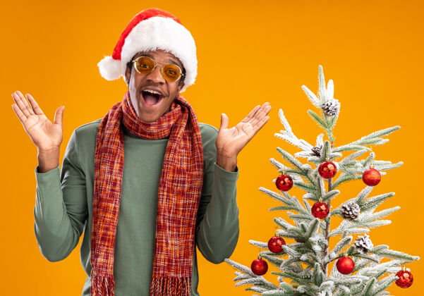 圣诞戴着圣诞帽 围着围巾的非洲裔美国人站在橙色背景下的圣诞树旁 开心地看着镜头男人周围相机