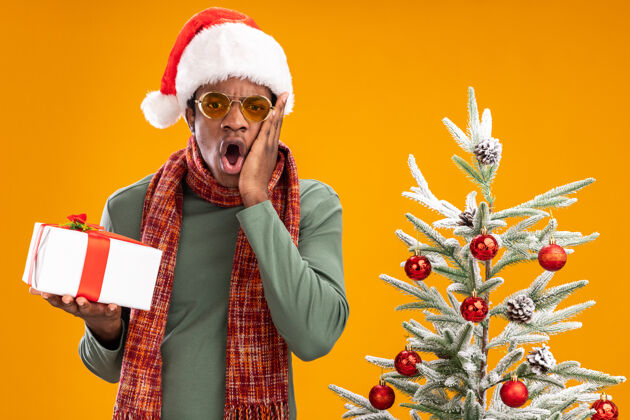 帽子一个戴着圣诞帽 脖子上围着围巾的非洲裔美国人拿着礼物 站在橙色背景下的圣诞树旁 看着相机 惊讶不已圣诞非洲礼物