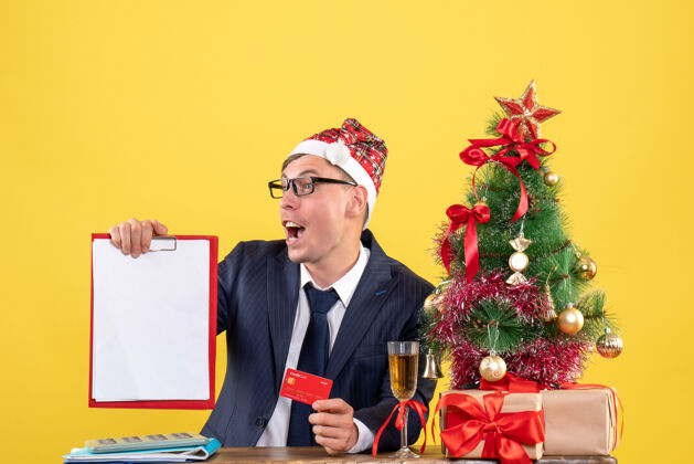 公司商务人士拿着剪贴板和卡片坐在圣诞树旁的桌子前 黄色的礼物剪贴板礼物职业