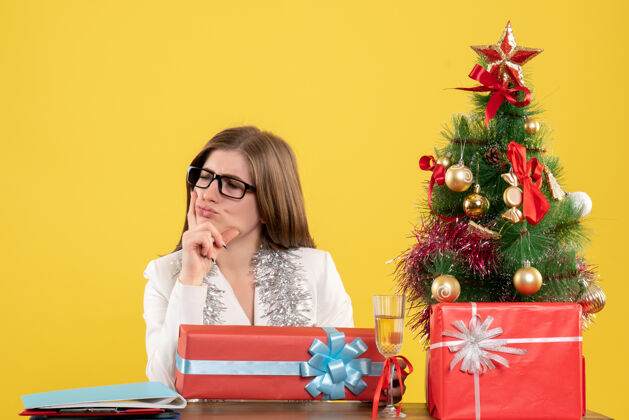 桌子前视图：女医生坐在桌子前 拿着礼物 黄色背景上的圣诞树和礼品盒女性礼物电脑