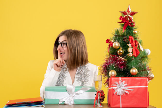 快乐正面图：女医生坐在桌子前 拿着礼物 黄色背景上的圣诞树和礼盒桌子树办公室