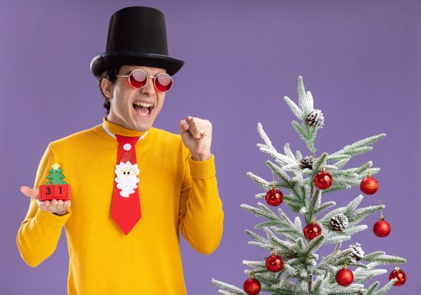 兴奋身穿黄色高领毛衣 戴着黑色帽子和滑稽领带的年轻人手里拿着新年约会的方块 兴奋而快乐地站在紫色背景下的圣诞树旁圣诞节年新的
