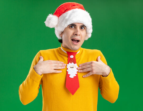 相机一个穿着黄色高领毛衣 戴着圣诞帽 打着滑稽领带的年轻人看着摄像机 惊讶地指着自己 双手站在绿色背景上高领毛衣站着手