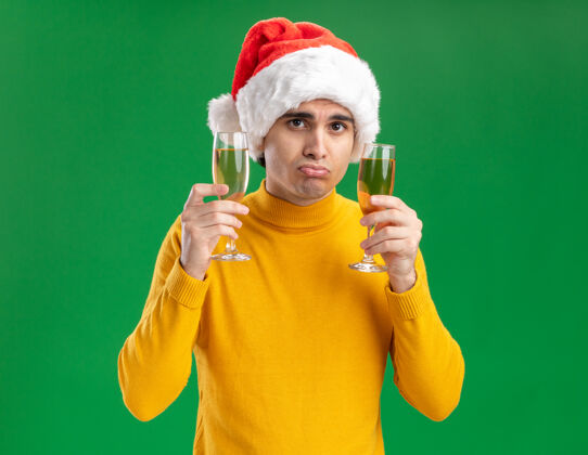 高领毛衣一个穿着黄色高领毛衣 戴着圣诞老人帽的年轻人 手里拿着两杯香槟 站在绿色的背景上 带着悲伤的表情看着镜头抱着帽子悲伤
