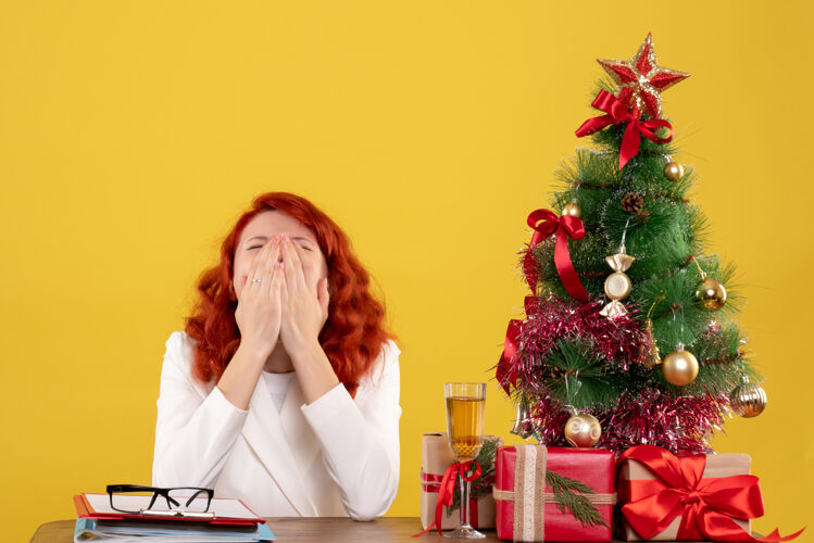 打哈欠前视图：女医生坐在桌子前 拿着礼物 圣诞树在黄色背景上打呵欠树新娘礼物