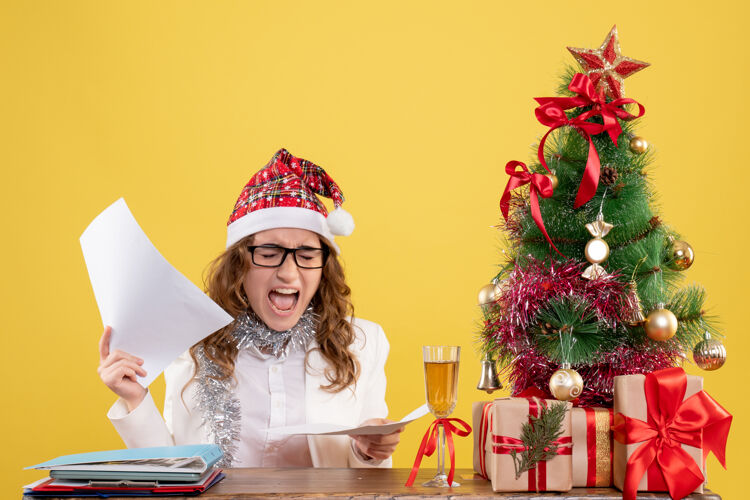 健康前视图女医生坐在圣诞树上 拿着黄色背景上的文件聚会礼物庆祝