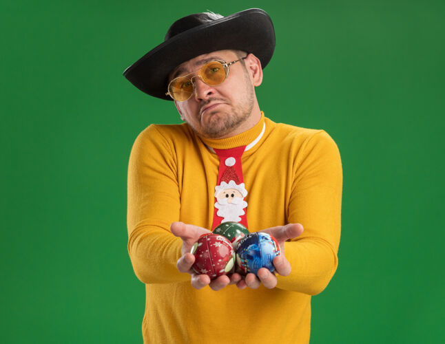 站着一个穿着黄色高领毛衣 戴着滑稽的红领带 戴着眼镜的年轻人手里拿着圣诞树玩具 脸上带着悲伤的表情站在绿色的墙上高领毛衣领带玩具