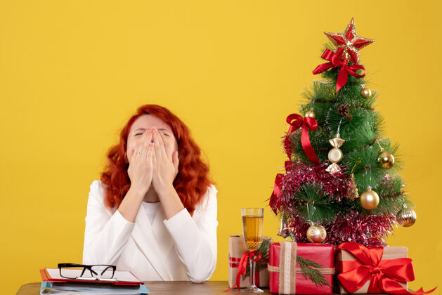 打哈欠前视图：女医生坐在桌子前 拿着礼物 圣诞树在黄色背景上打呵欠树新娘礼物