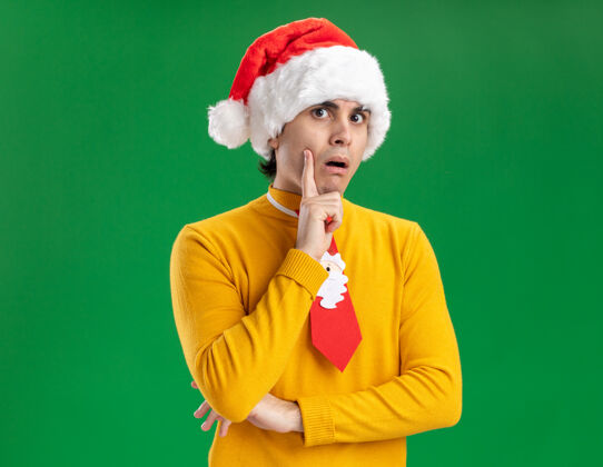 帽子一个穿着黄色高领毛衣 戴着圣诞老人帽 打着滑稽领带的年轻人站在绿色背景下 困惑而担忧地看着摄像机年轻看相机