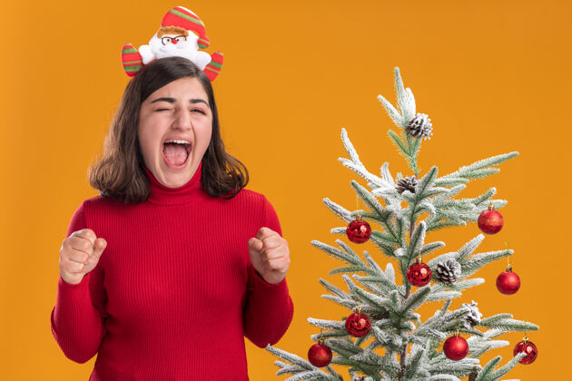 年轻穿着圣诞毛衣的年轻女孩戴着有趣的头带 在橙色背景下的圣诞树旁快乐而兴奋拳头毛衣疯狂