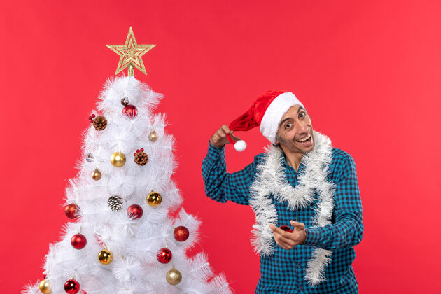 帽子快乐的年轻人在玩圣诞老人的帽子在蓝色的衬衫和圣诞树上的红色庆祝玩男人