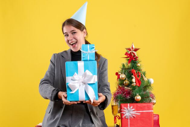 近正面图：戴着派对帽的欢欣女孩双手捧着圣诞礼物 靠近圣诞树和礼物鸡尾酒桌子高兴成人