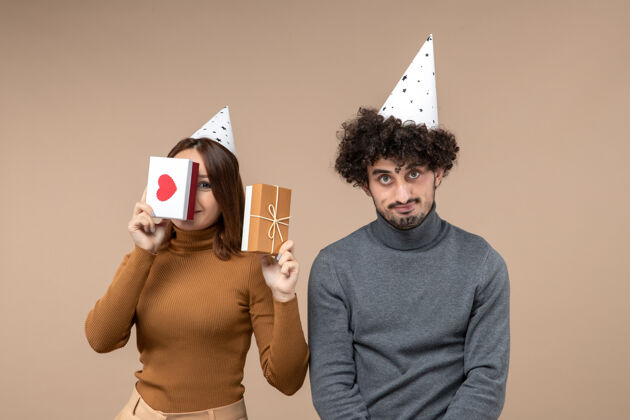 年轻的情侣新年概念与年轻夫妇戴新年帽女孩关闭她的脸心和礼物和不满意的家伙礼物伪装面具
