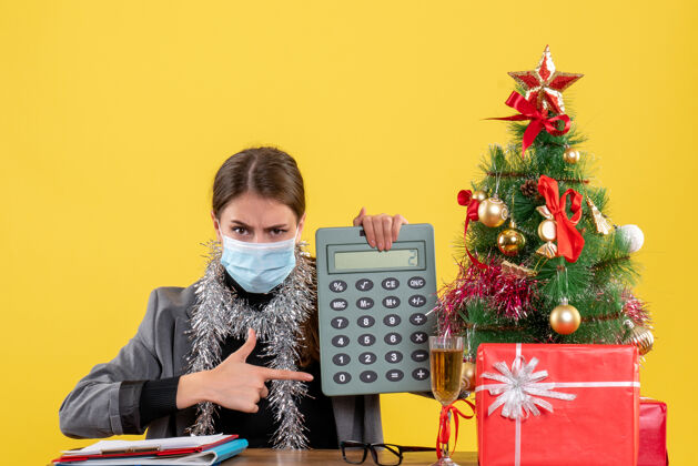 计算器正面图严肃的年轻女孩戴着医用口罩坐在桌边用手指指着计算器圣诞树和礼物鸡尾酒广播磁盘赛马磁盘