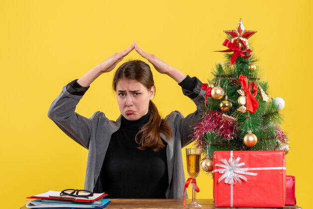 树前视图悲伤的女孩坐在书桌旁 手举过头顶 像一个屋顶房子靠近圣诞树和礼物鸡尾酒头礼物女孩