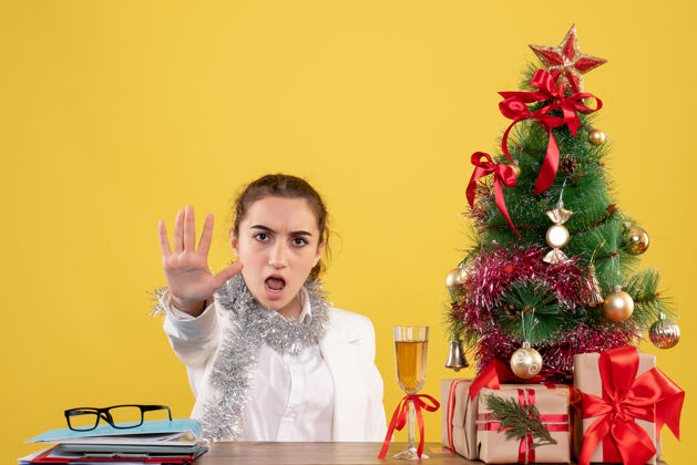 背景前视图：女医生坐在黄色背景的桌子后面 带着圣诞树和礼品盒办公室前面桌子