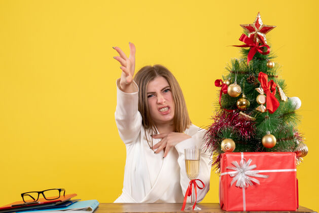 圣诞节前视图女医生坐在她的桌子前 不高兴的黄色背景与圣诞树和礼品盒医生不高兴桌子