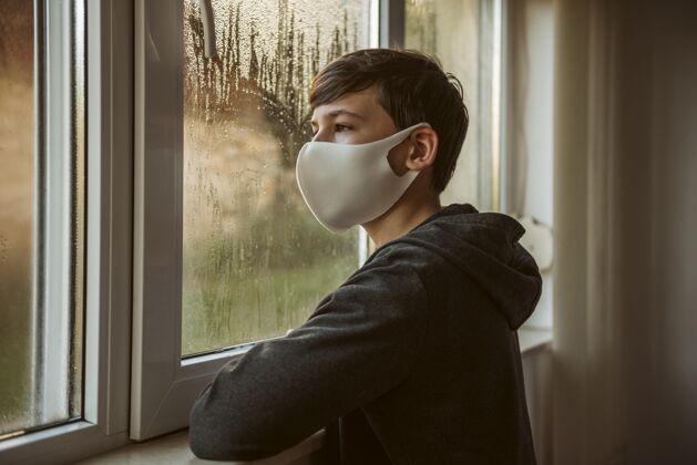 冠状病毒戴着面罩的男孩从窗户往外看青少年大流行隔离