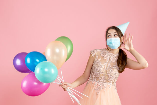 粉红色戴着派对帽和医用面具的快乐女孩一边听着 一边拿着粉色的彩色气球气球听聚会